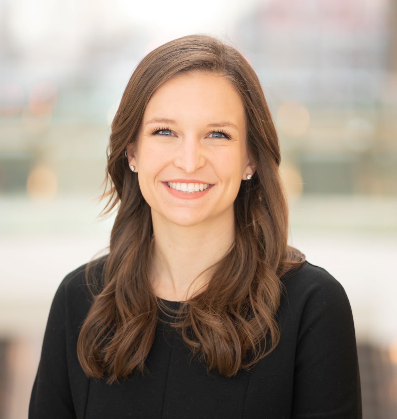 A smiling woman with brown hair and blue eyes, wearing pearl earrings and a black top, in an indoor setting with blurred background.