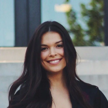 A young woman with long dark hair smiling outside with a modern building and green trees in the background.
