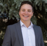 A professional woman smiling outdoors with trees in the background, wearing a blazer and white shirt.