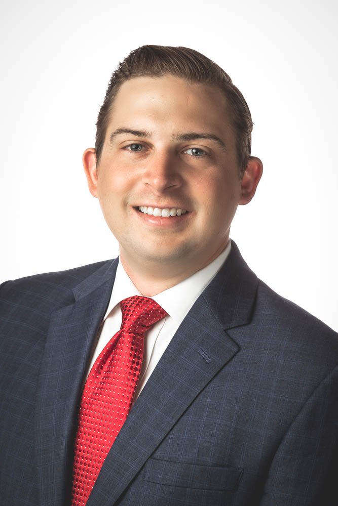 A professional portrait of a young man with fair skin, wearing a dark suit with a subtle check pattern, a white shirt, and a red tie, smiling against a plain white background.