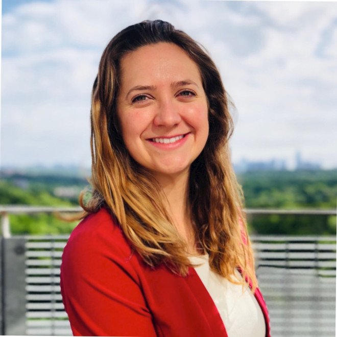 Smiling woman with long wavy hair wearing a red blazer and white top, outdoors with a cloudy sky and distant cityscape in the background.