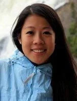 A young woman with long dark hair smiling outdoors, wearing a light blue rain jacket.