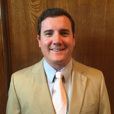 A man with dark hair wearing a tan suit, white shirt, and a light-colored tie, smiling in front of a wooden wall.
