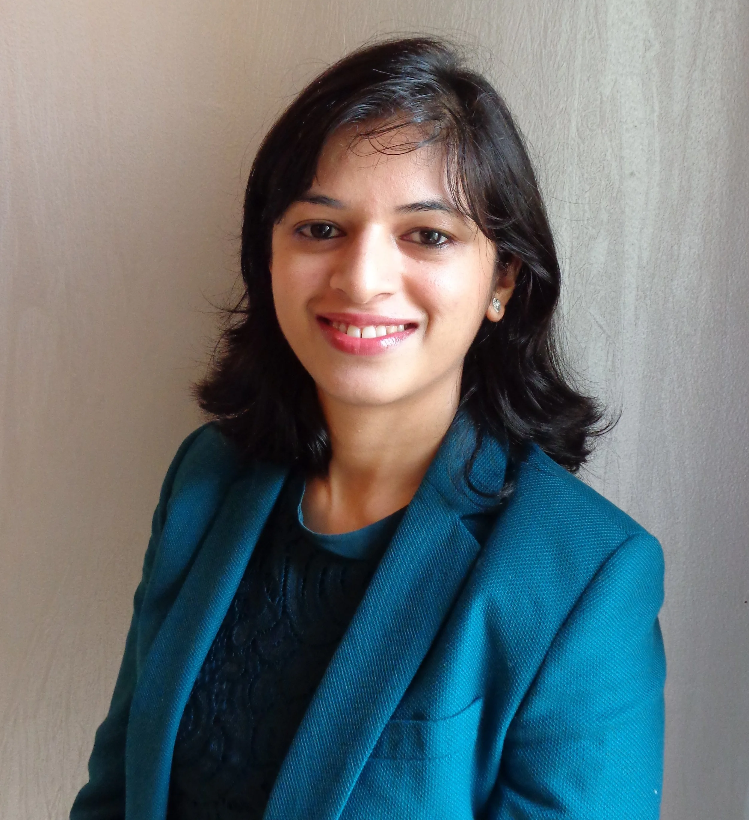 A woman with shoulder-length black hair wearing a teal blazer and matching top, smiling in front of a plain wall.