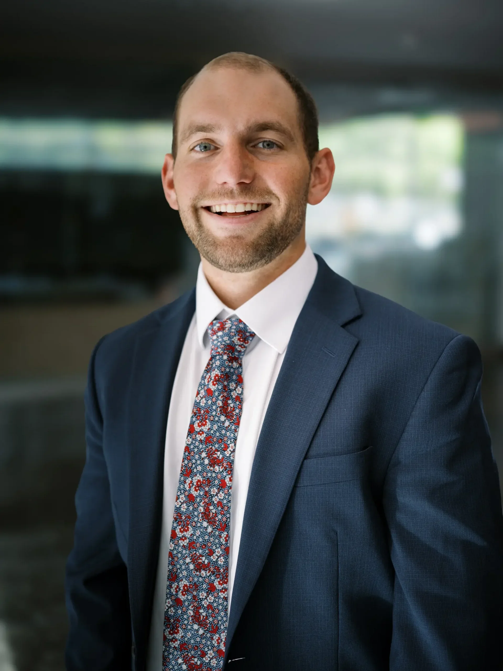 A smiling man in a navy suit, white shirt, and a colorful patterned tie, posing in an indoor setting with a blurred background.