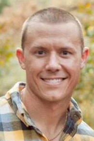 A smiling man with short hair wearing a plaid shirt, outdoors with a blurred autumn background.