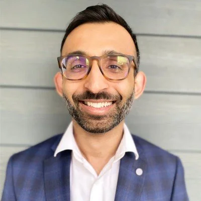 A smiling man with glasses, a beard, wearing a blue checkered blazer and a white shirt, standing in front of a gray wooden wall.