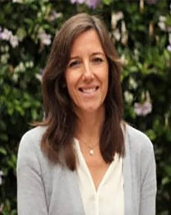 A woman with shoulder-length brown hair smiling outdoors in front of green foliage.