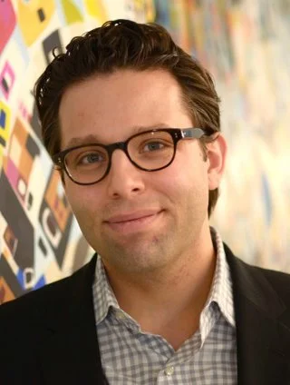 A man with glasses and dark hair, wearing a checkered shirt and a dark blazer, smiling in front of a colorful, patterned background.