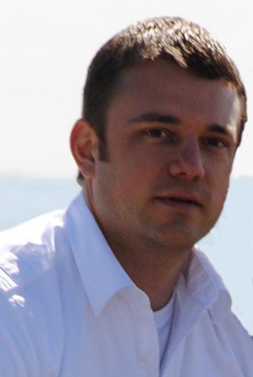 A young man with short dark hair wearing a white collared shirt outdoors with a bright sky and water in the background.