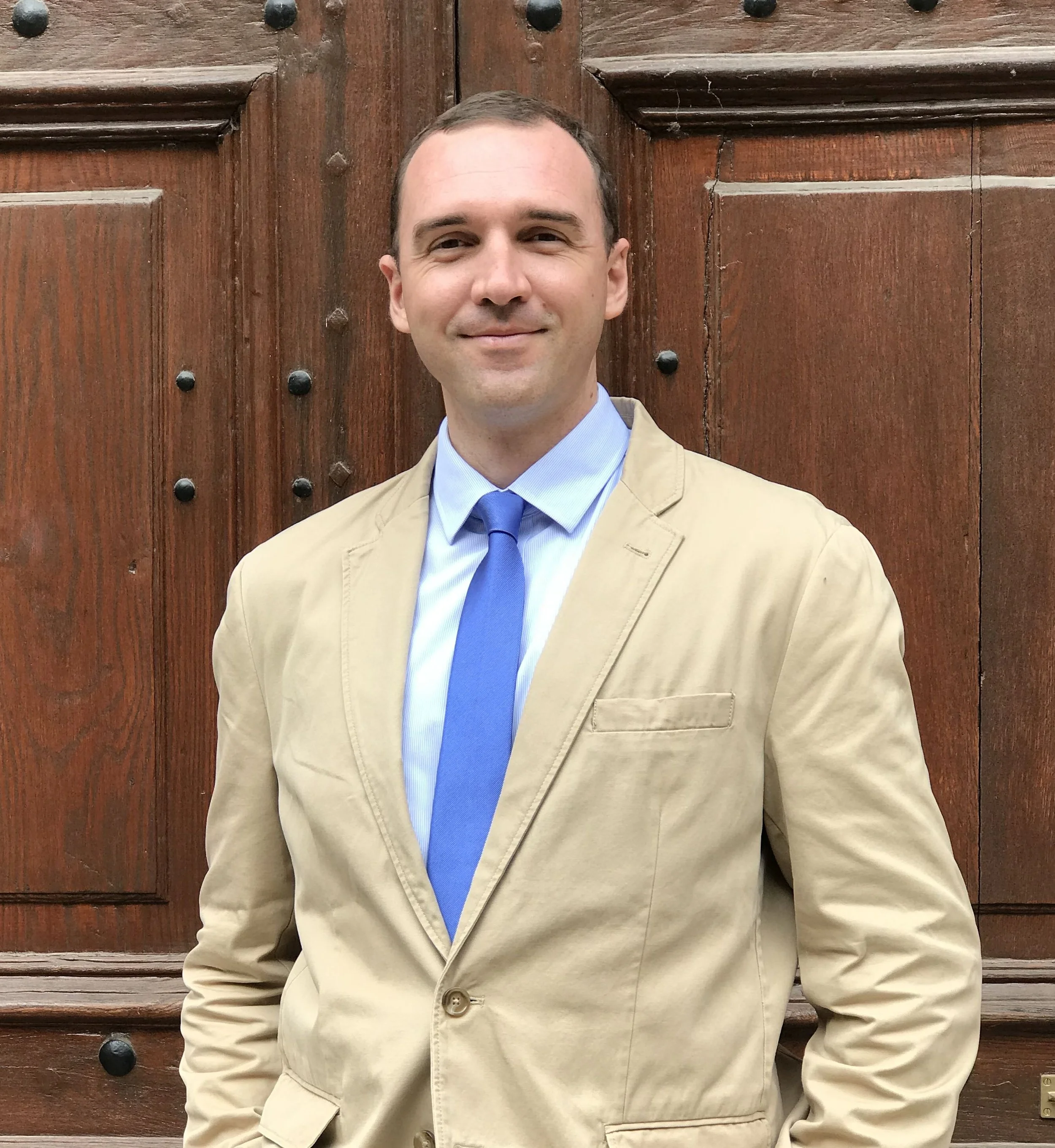A man in a beige blazer, light blue shirt, and blue tie standing in front of a brown wooden door with black iron studs.