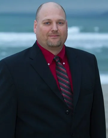 A man in a dark suit, red shirt, and striped tie standing on a beach with the ocean and waves in the background.