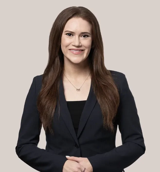 A woman with long brown hair wearing a black blazer and a necklace, smiling at the camera against a plain background.