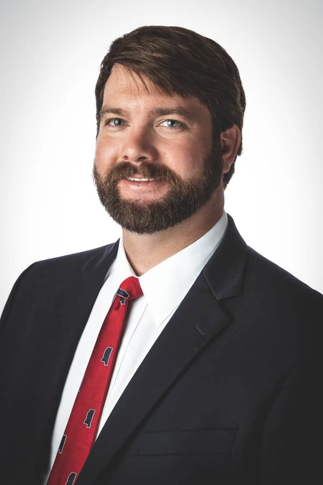 Professional headshot of a man with dark hair and beard, wearing a black suit, white shirt, and red tie, smiling against a plain light gray background.