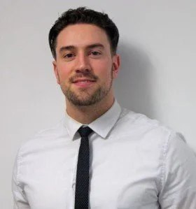 A young man with dark hair and a beard, wearing a white shirt and black tie, smiling against a plain background.