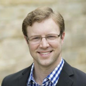 A man with glasses, light brown hair, and a light complexion, smiling in front of a blurred outdoor background, dressed in a dark blazer and checked shirt.