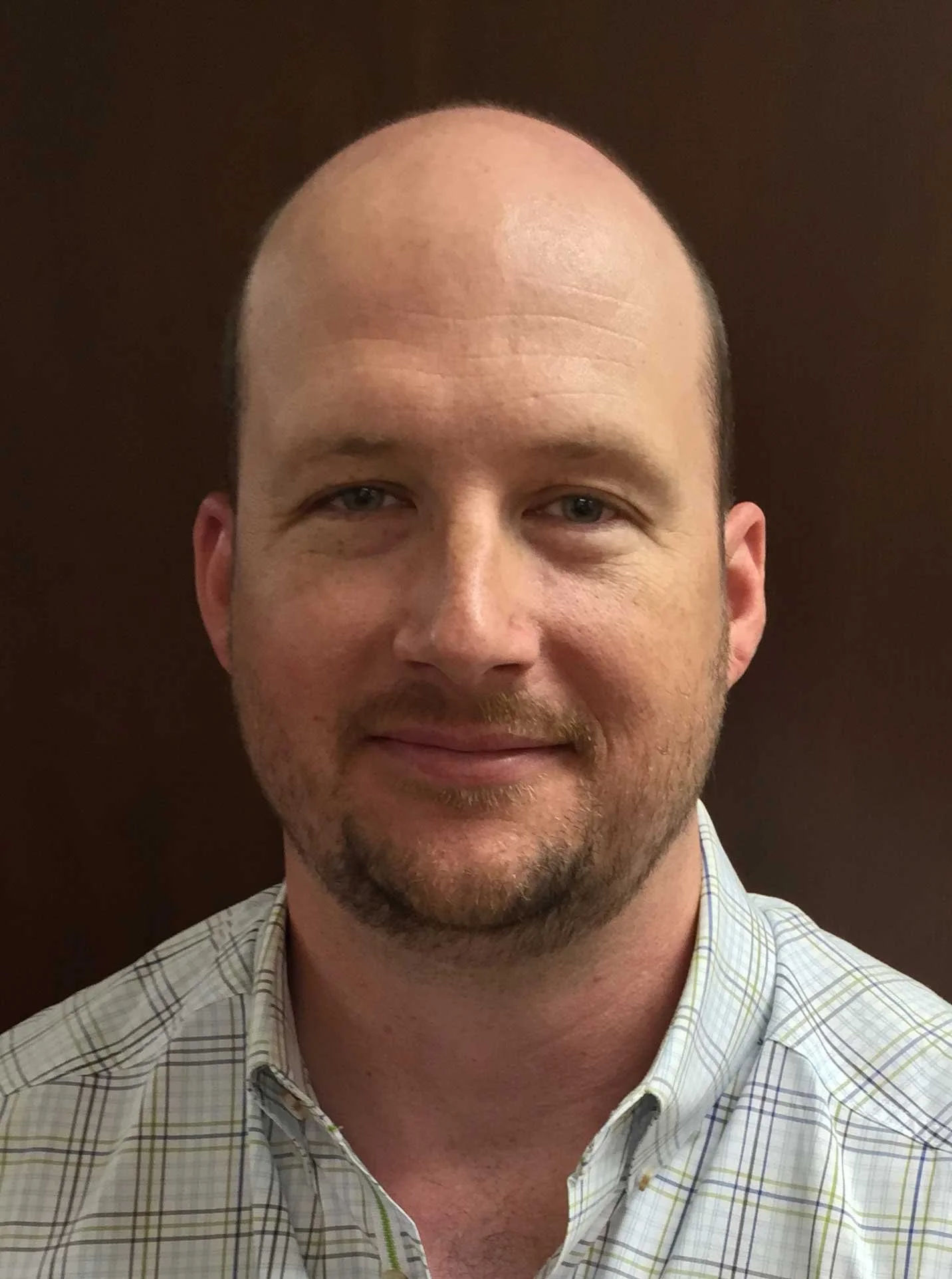 Portrait of a middle-aged man with short hair and a beard, wearing a checkered shirt, standing against a dark background.