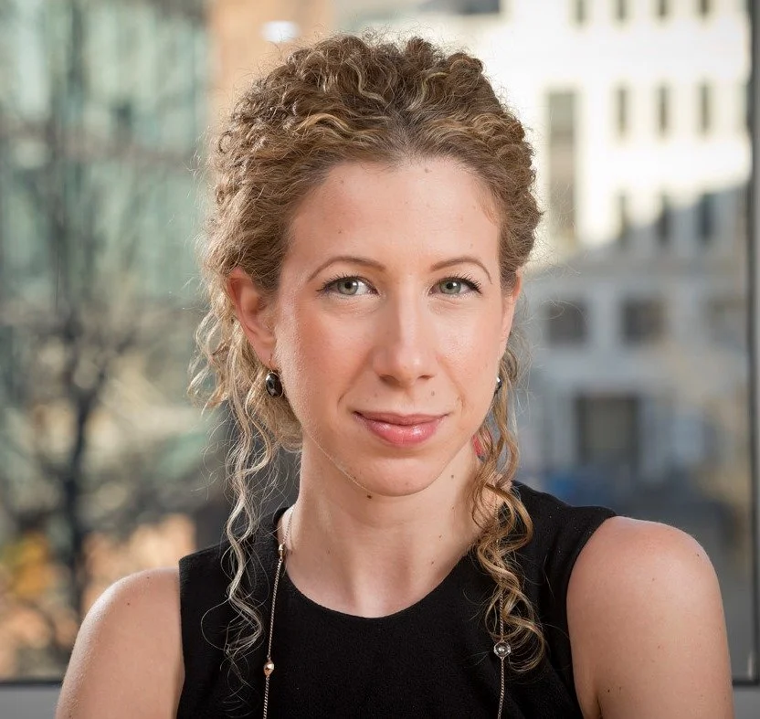 A woman with curly blonde hair, wearing a black sleeveless top, sitting in front of a window with a cityscape background.
