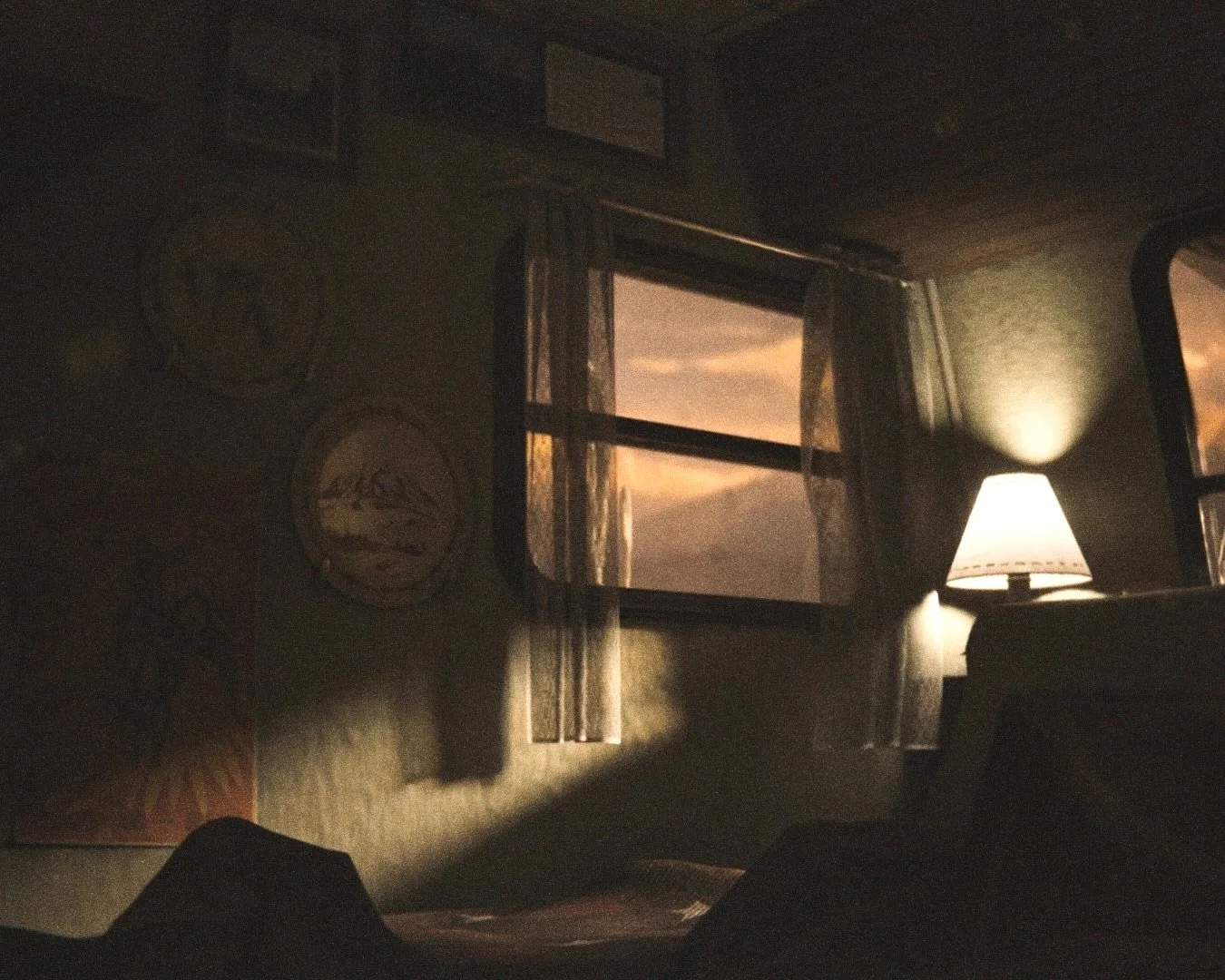 A dimly lit room with a window showing a sunset sky, decorated with framed pictures and circular wall hangings, illuminated by a table lamp.