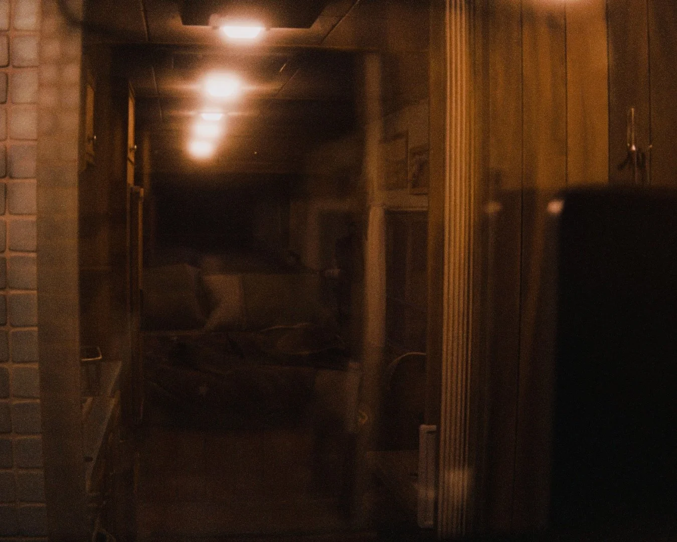 Dimly lit room with wooden walls, a chair, and a few shelves or cabinets, viewed through a reflective glass or window.