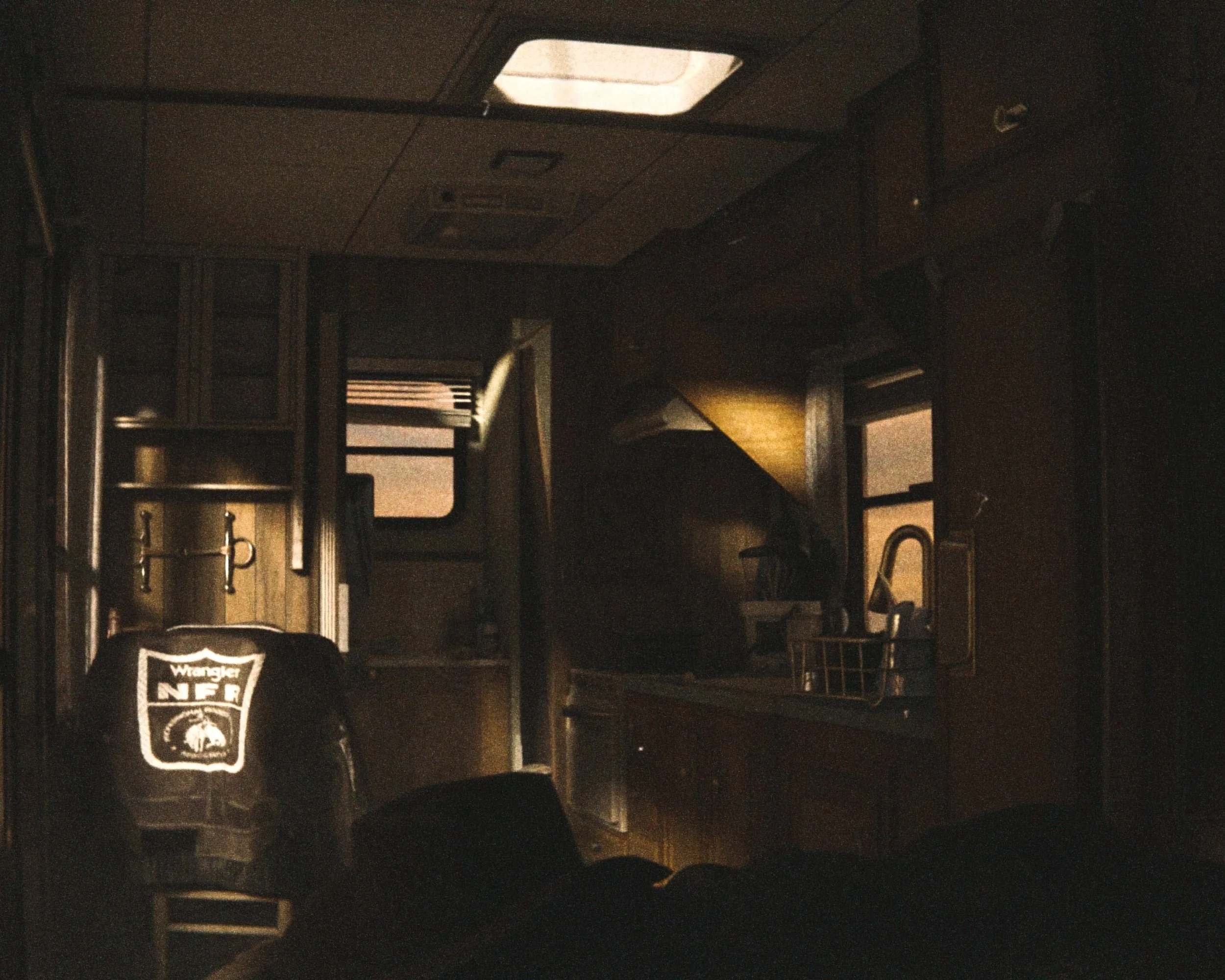 Dark interior of a bus or RV with windows on each side and furniture, including tables and seats. Overcast light coming through the windows, with a sports backpack in the foreground that has a Black Raiders logo.