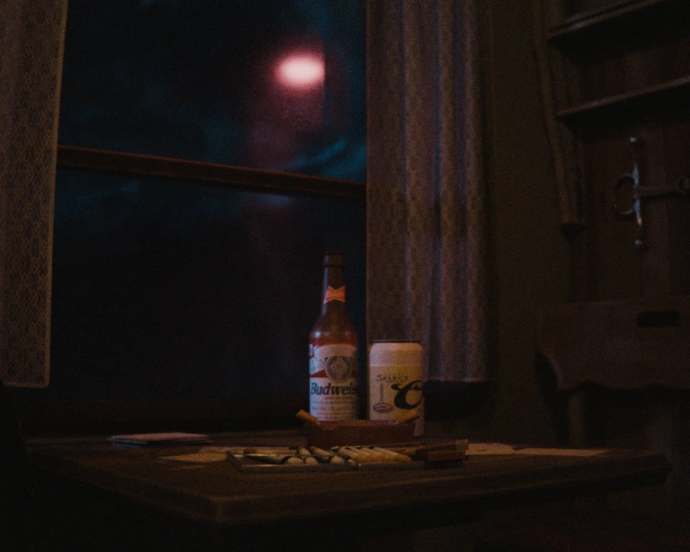 Dimly lit scene with a Budweiser beer bottle, a disposable coffee cup, and a cigar on a tray on a wooden table, with a window showing an outdoor night sky with the moon.