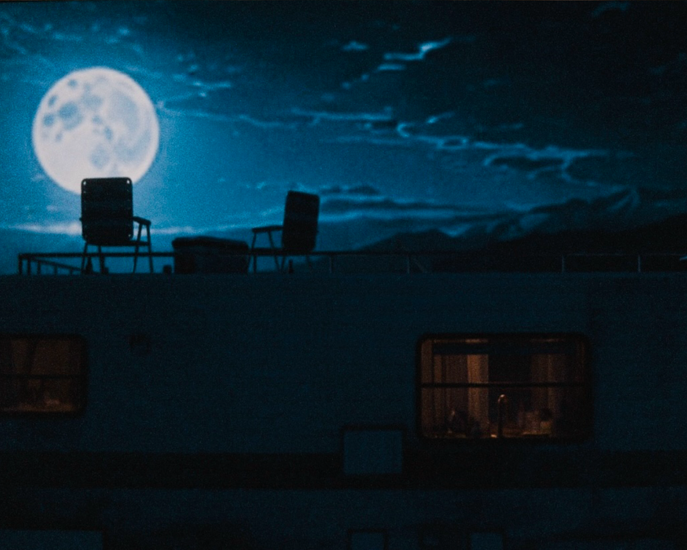 Nighttime scene of a building with two chairs and a small table on the rooftop, illuminated by the full moon and starry sky in the background.
