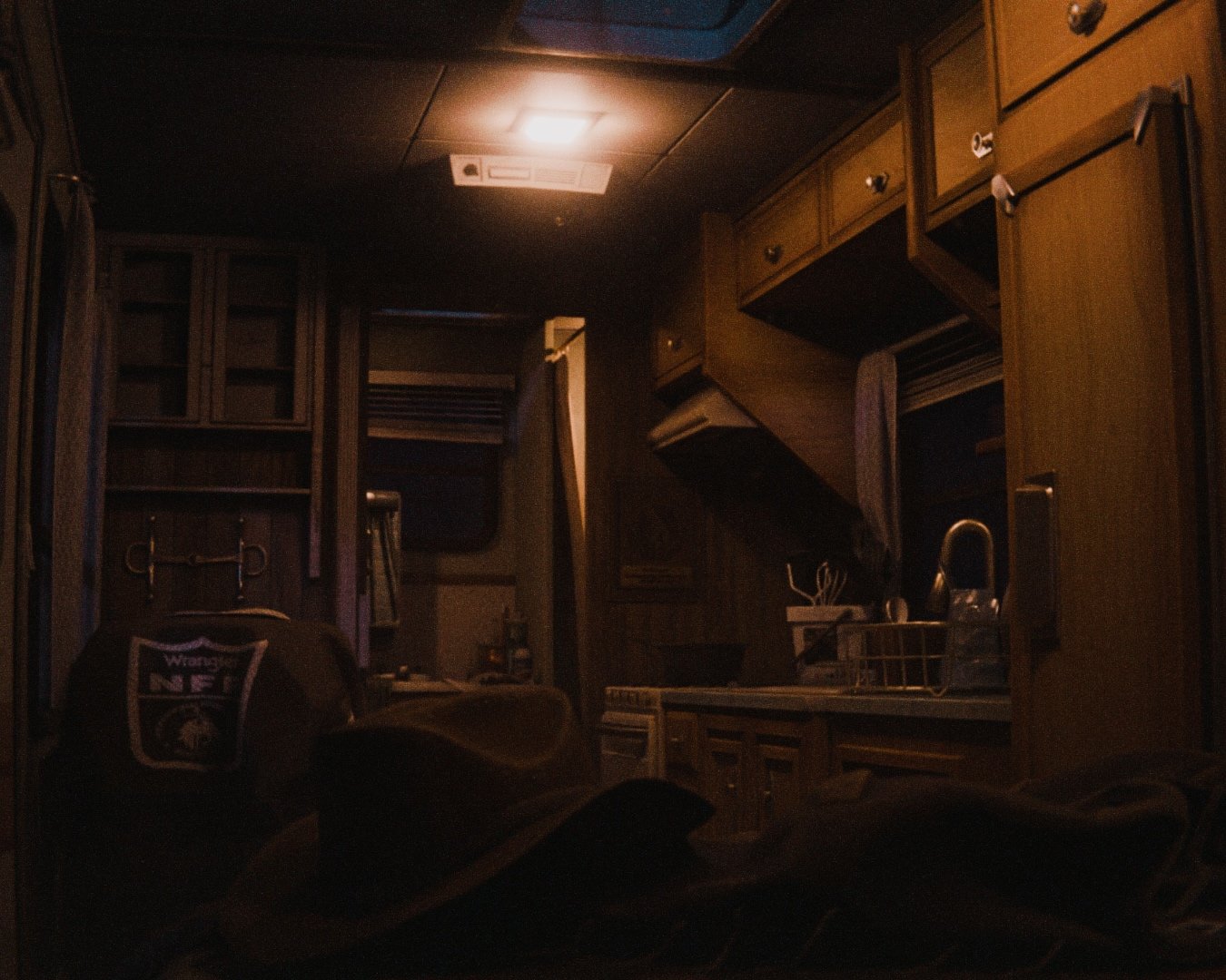 Dark, dimly lit kitchen with wooden cabinets, a sink with a faucet, a dish rack, and a countertop with various items. A person wearing a jacket with a 'Warrenville NF' logo is sitting in the foreground.