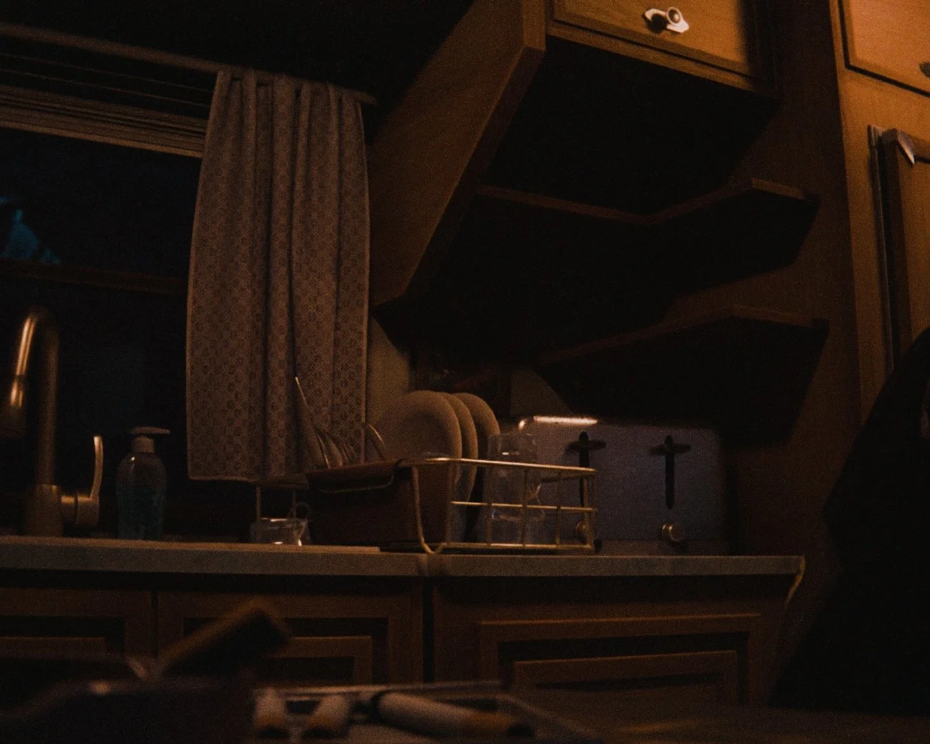 Dark kitchen scene with dishes drying on the counter, a window with curtains, and kitchen appliances including a toaster and a bottle of soap.