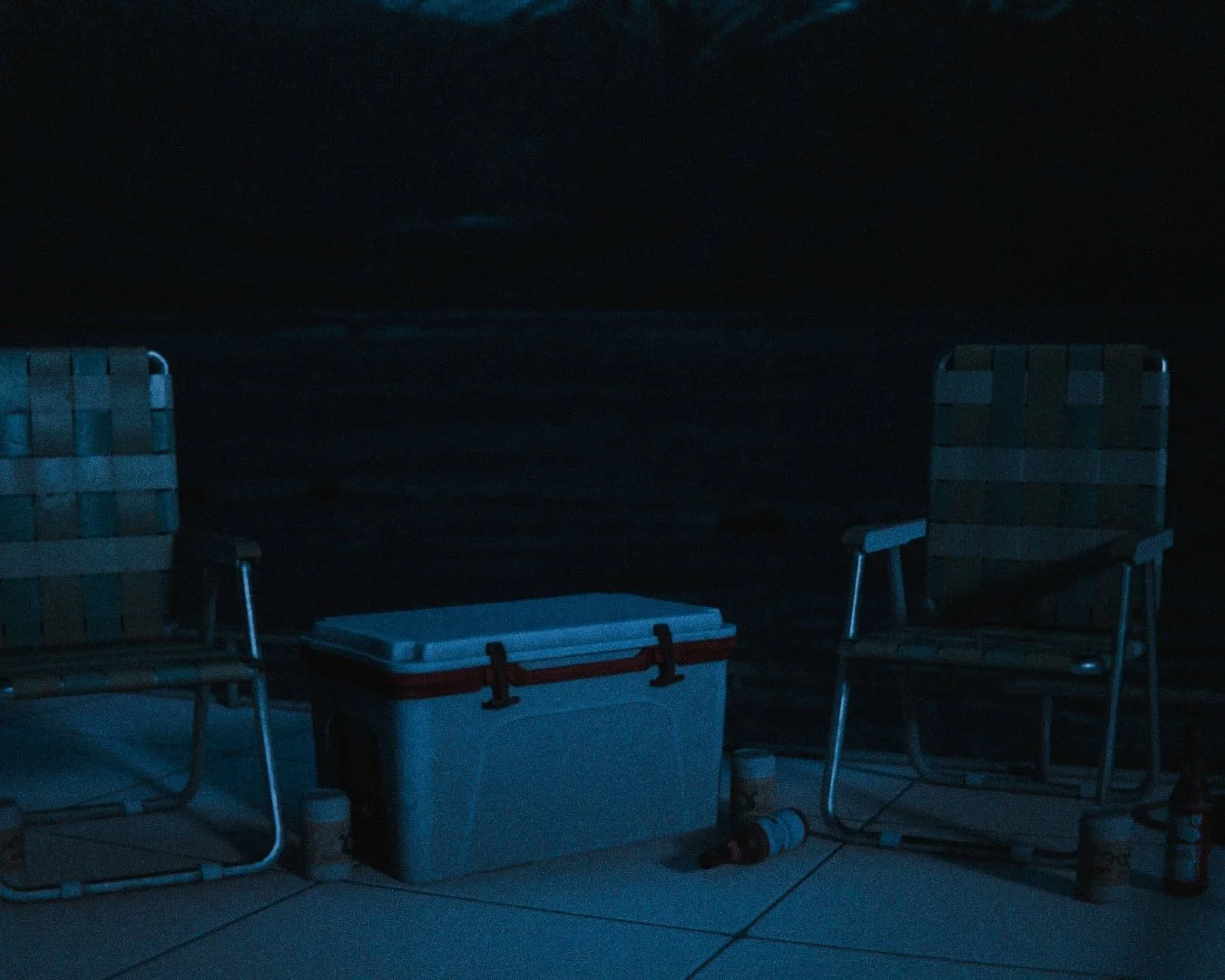 Two folding chairs, a cooler, and some bottles placed on a concrete deck near a dark beach or water, with the night sky overhead.