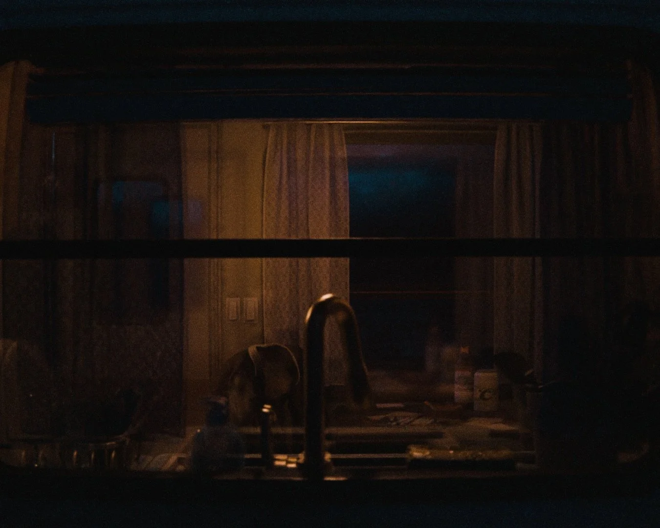 A dimly lit kitchen viewed through a glass window or door, with a sink, faucet, and various kitchen items visible in the foreground, and a room with curtains and a television or monitor in the background.