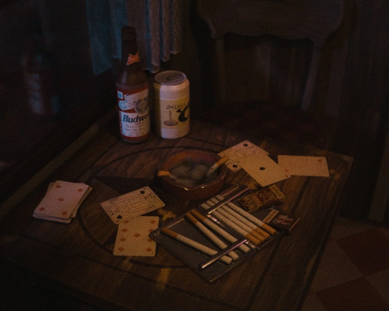 A wooden table with a pack of cigarettes, a pack of matches, an ashtray with cigarette butts, and playing cards scattered on it, with bottles of beer and whiskey on a nearby surface.