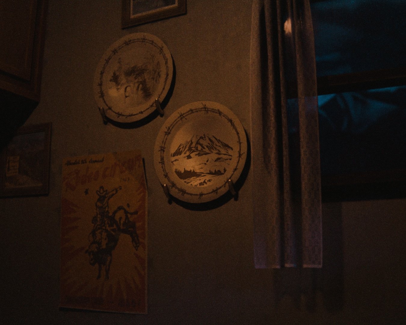 Dimly lit room with art pieces on the wall, including two circular wooden engravings with mountain and landscape designs, and a poster depicting a cowboy figure on a rearing horse with a bright background. Wood paneling partly visible on the right.