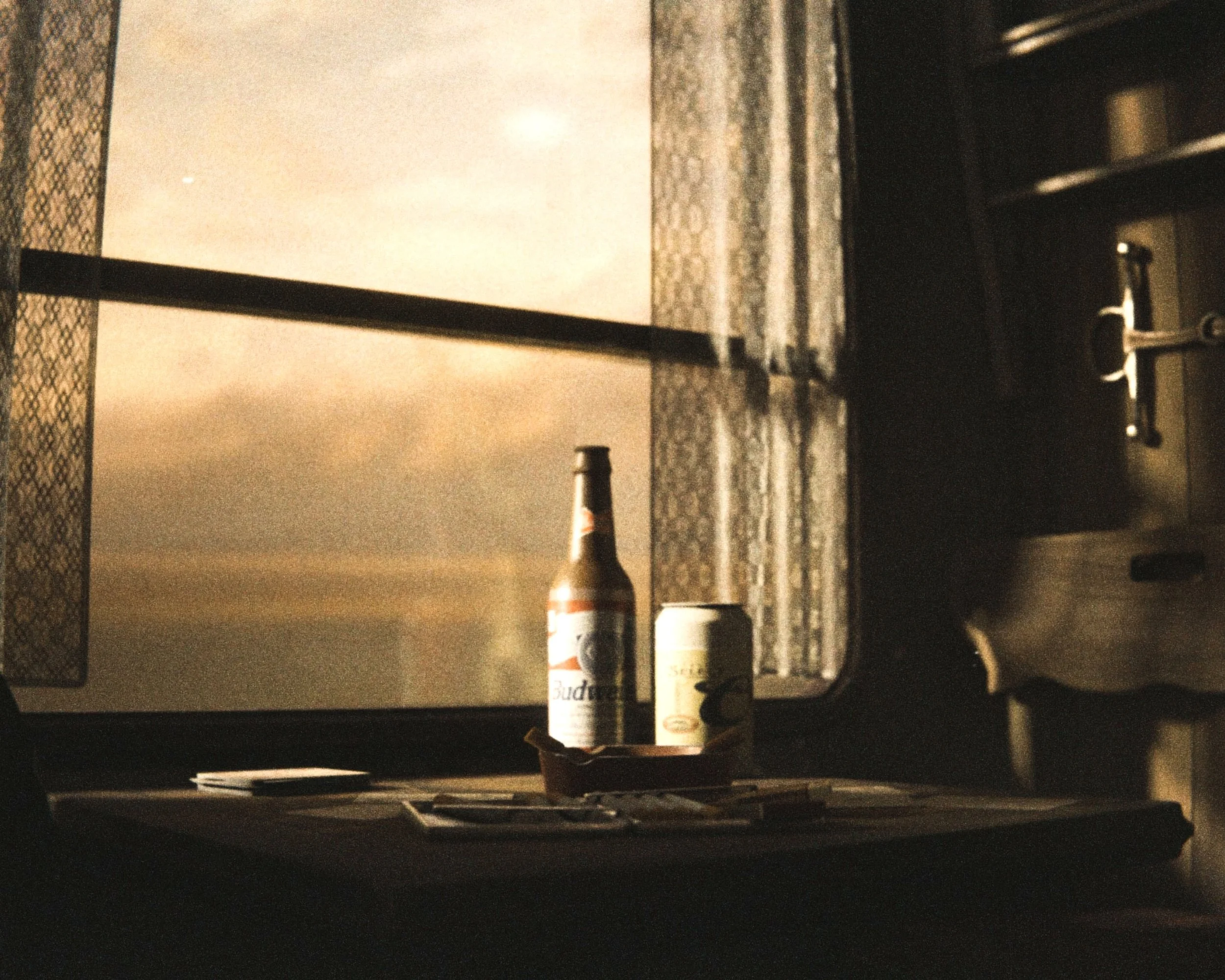 A dimly lit dinner table with a bottle of Budweiser beer, a salt shaker, and a yellow mustard bottle in the foreground. In the background, there is a window with patterned curtains and a patterned surface on the table.