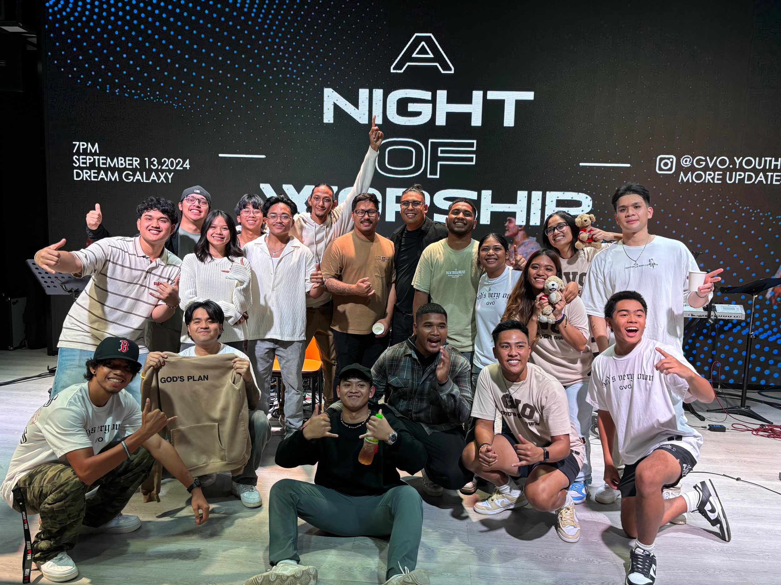 A group of people smiling and posing on stage at an event. The backdrop displays the words 'A Night of Worship' with the date September 13, 2024, at Dream Galaxy. The group includes both men and women, some wearing casual clothing, with one person ho