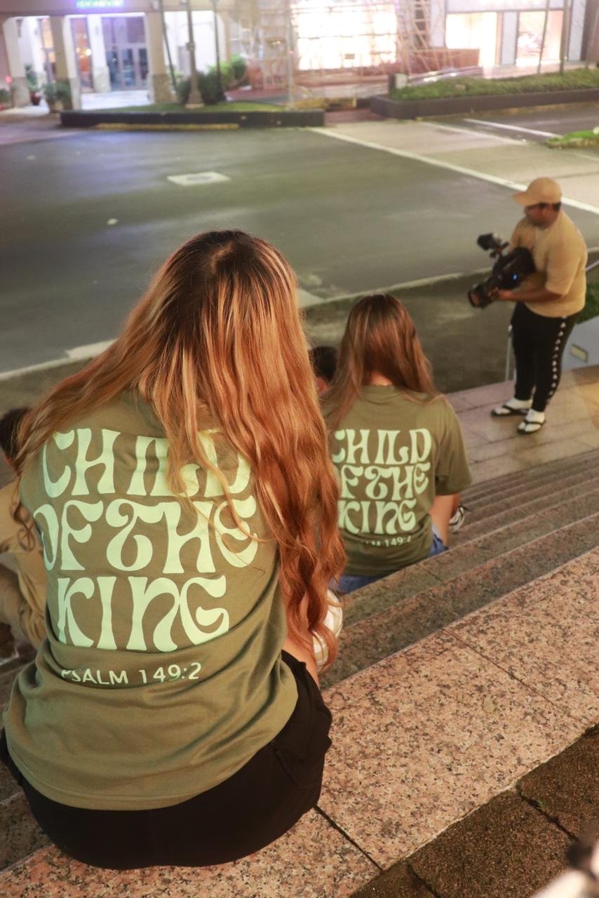 Group of people sitting on stairs, wearing matching T-shirts with 'Child of the King - Psalm 149:2' printed on the back, a person on the side holding a camera.