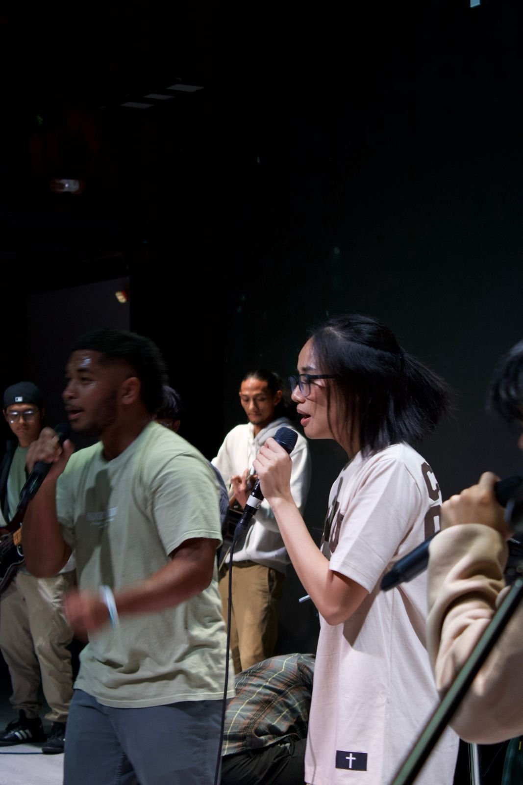 Group of young musicians performing on stage, with two individuals singing into microphones and others playing instruments in the background.