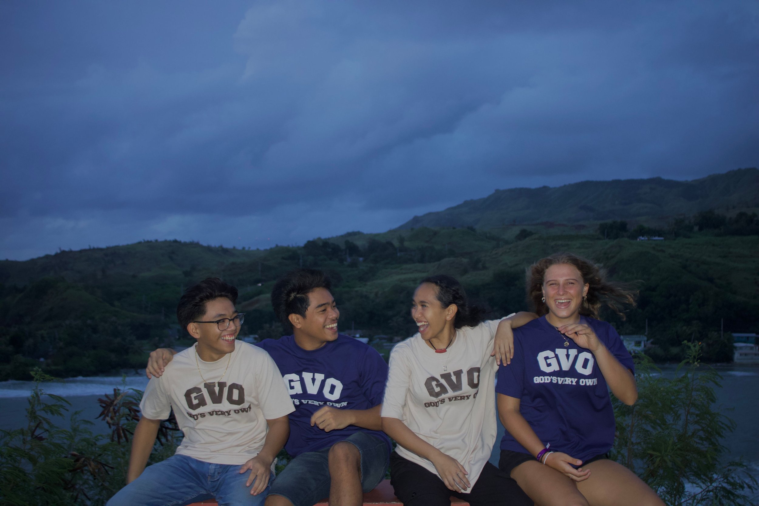 Four friends sitting outdoors, smiling and wearing "GVO God's Very Own" t-shirts, with a scenic background of hills and a body of water at dusk.