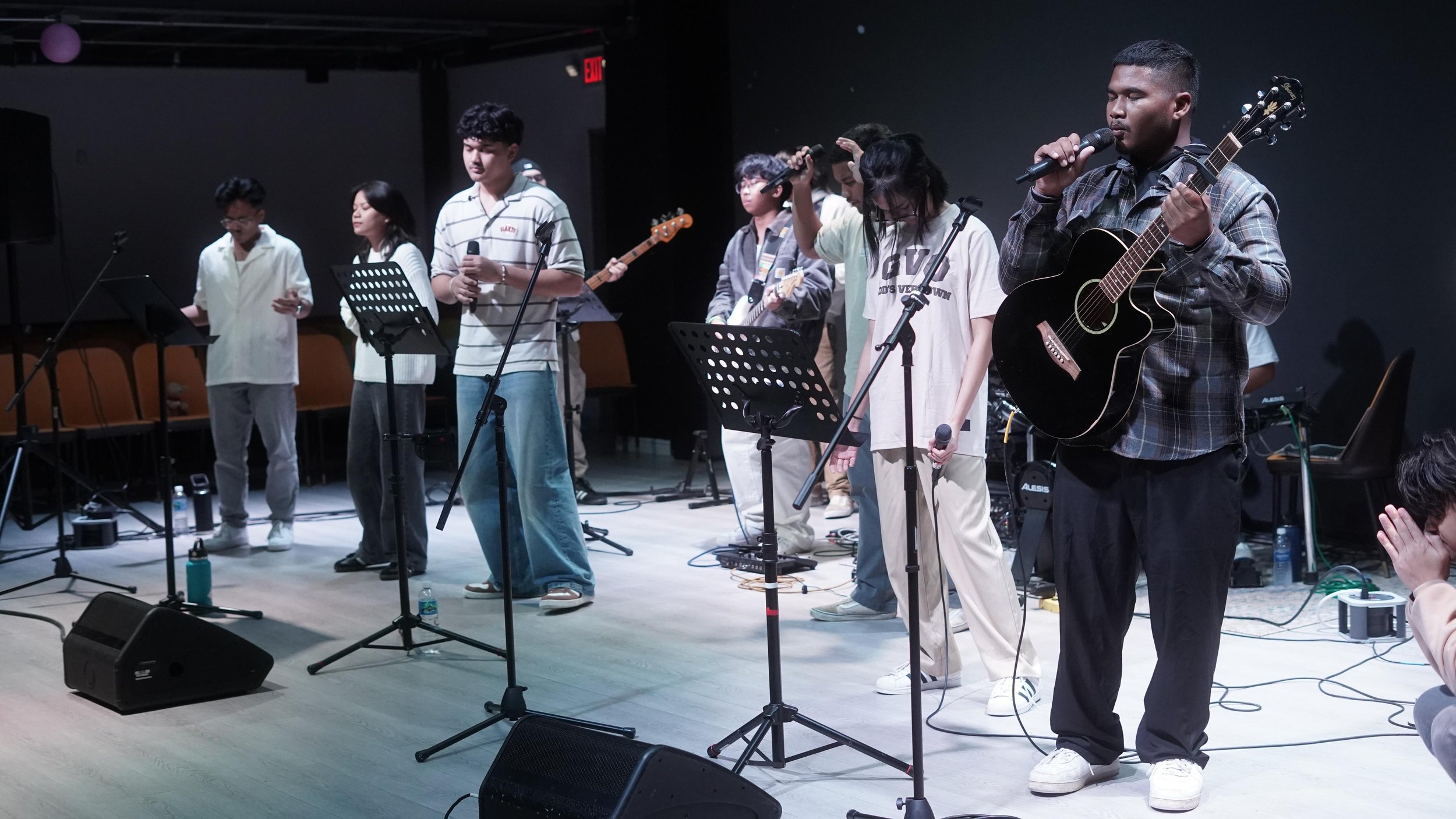 A group of musicians and singers perform on stage, with one person holding a black acoustic guitar and a microphone. Other members are holding microphones and music stands are present. The lighting is focused on the performers.