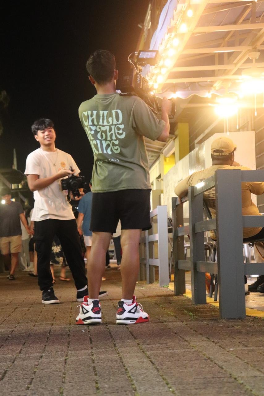 Two men with cameras filming outside a lit building at night, one wearing a shirt with "Child of the King" on the back.
