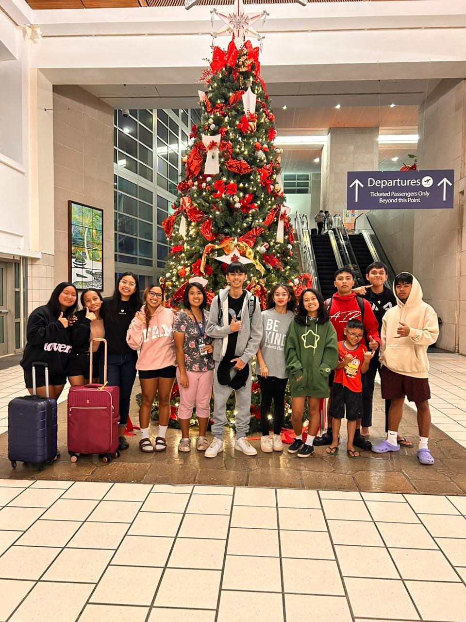 A group of people posing in front of a decorated Christmas tree in an airport terminal. They stand on a tiled floor, with suitcases nearby. The background shows signs for departures and an escalator.