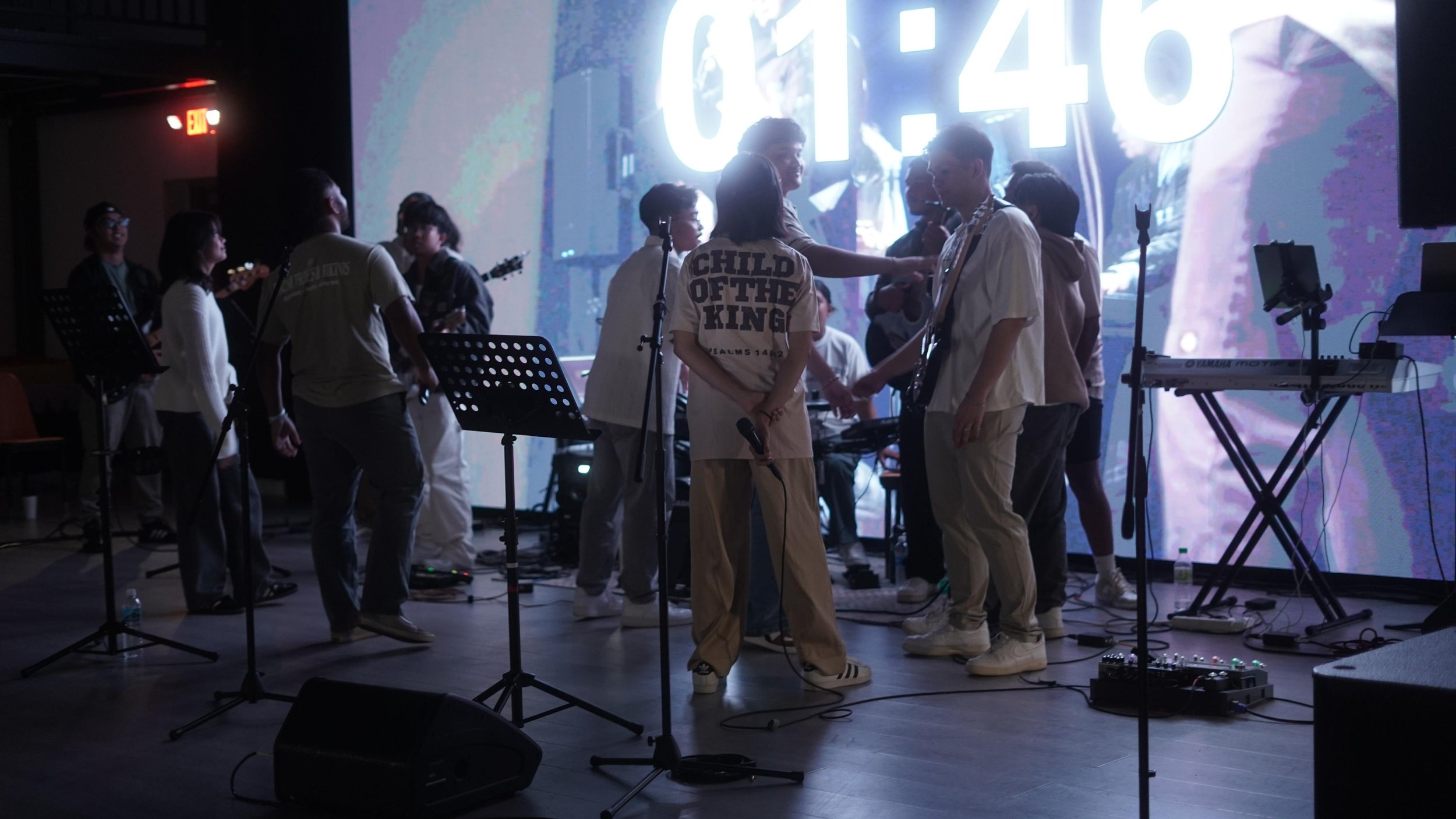 Group of people on stage with musical equipment, microphones, and a countdown timer displayed on a screen in the background.