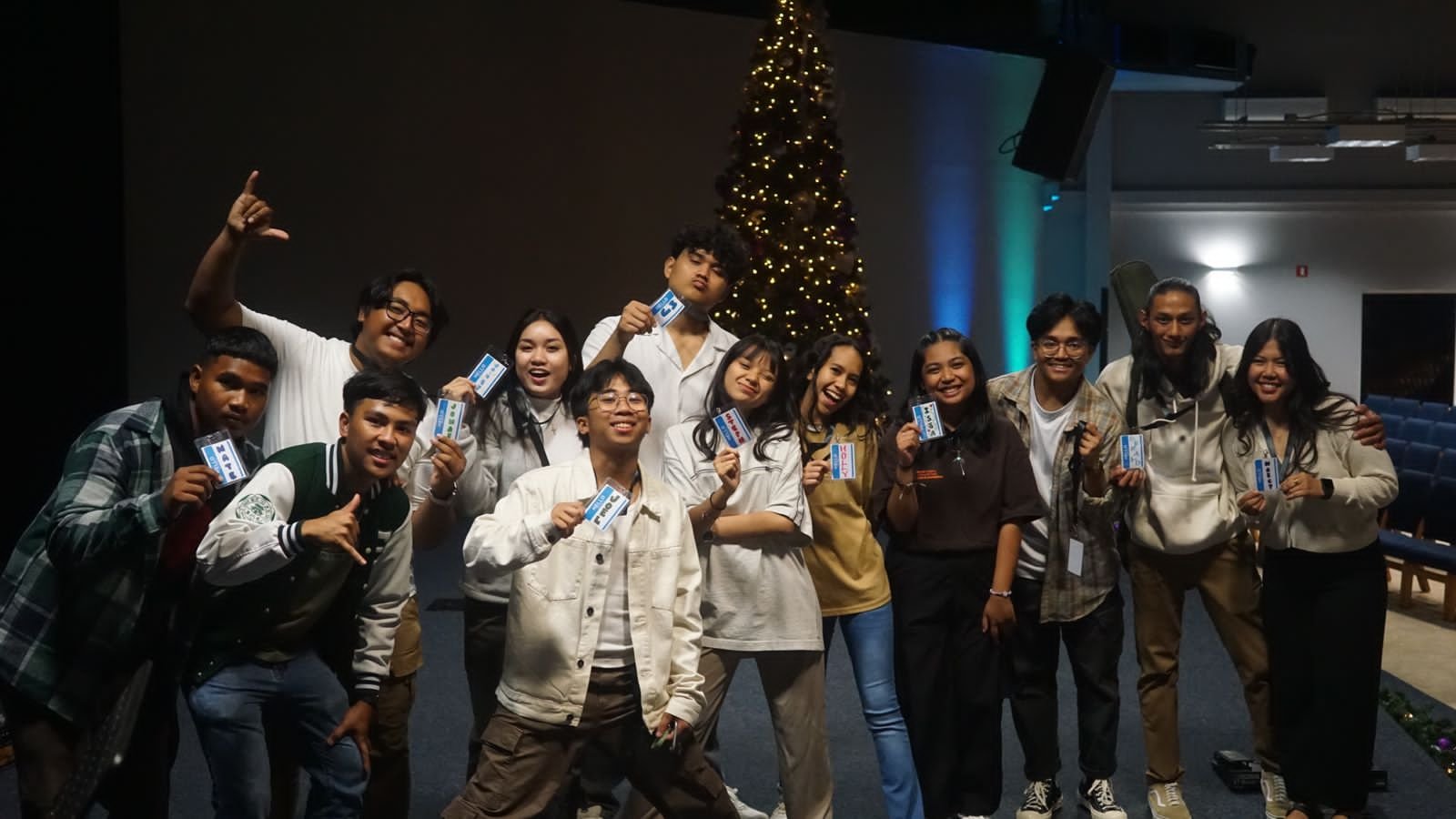Group of diverse young adults smiling and holding cards, standing in front of a decorated Christmas tree indoors.