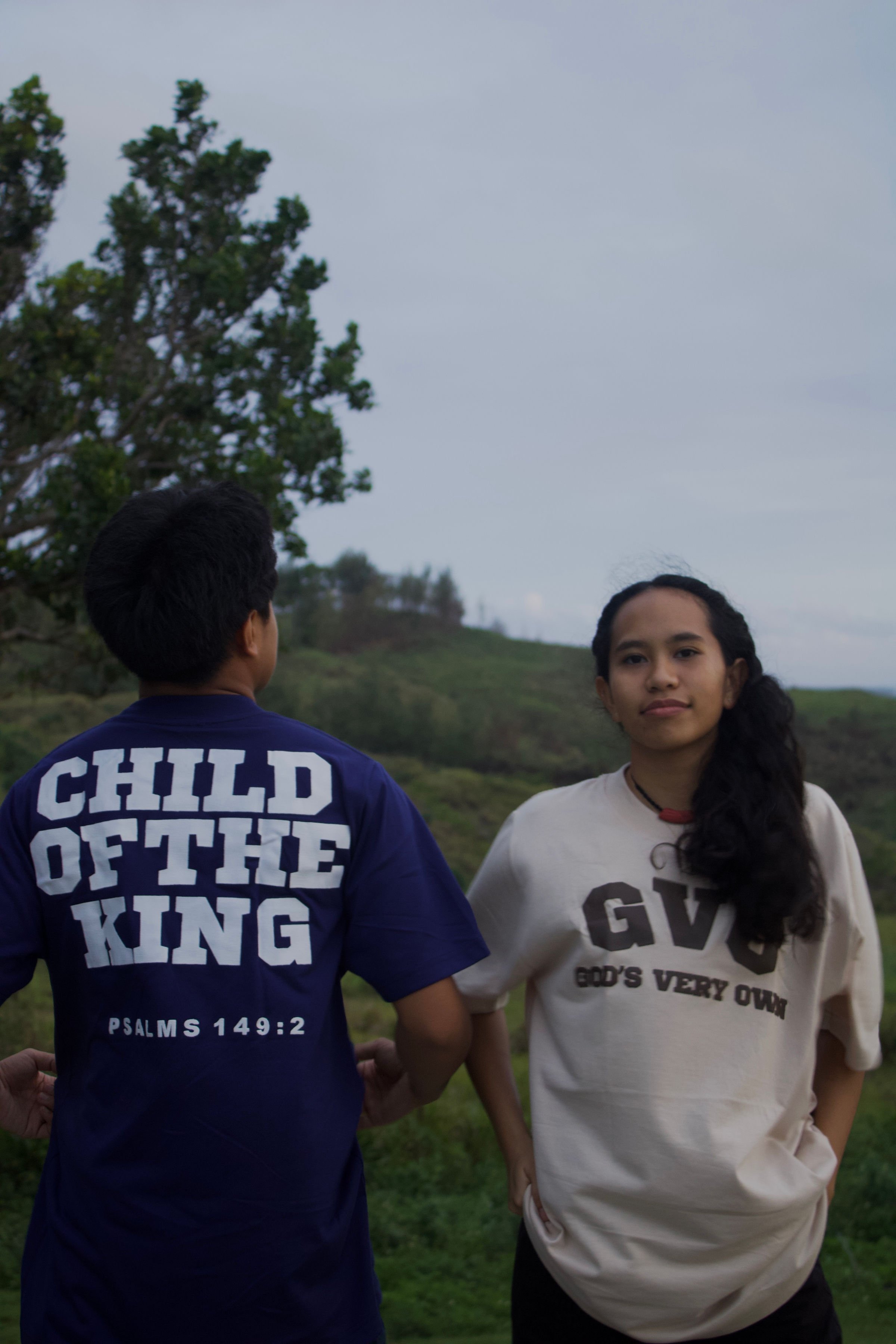 Two individuals wearing t-shirts with religious messages. The person on the left, facing away, has "Child of the King - Psalms 149:2" on their shirt. The person on the right, facing forward, wears a shirt with "GVO - God's Very Own." They are standin