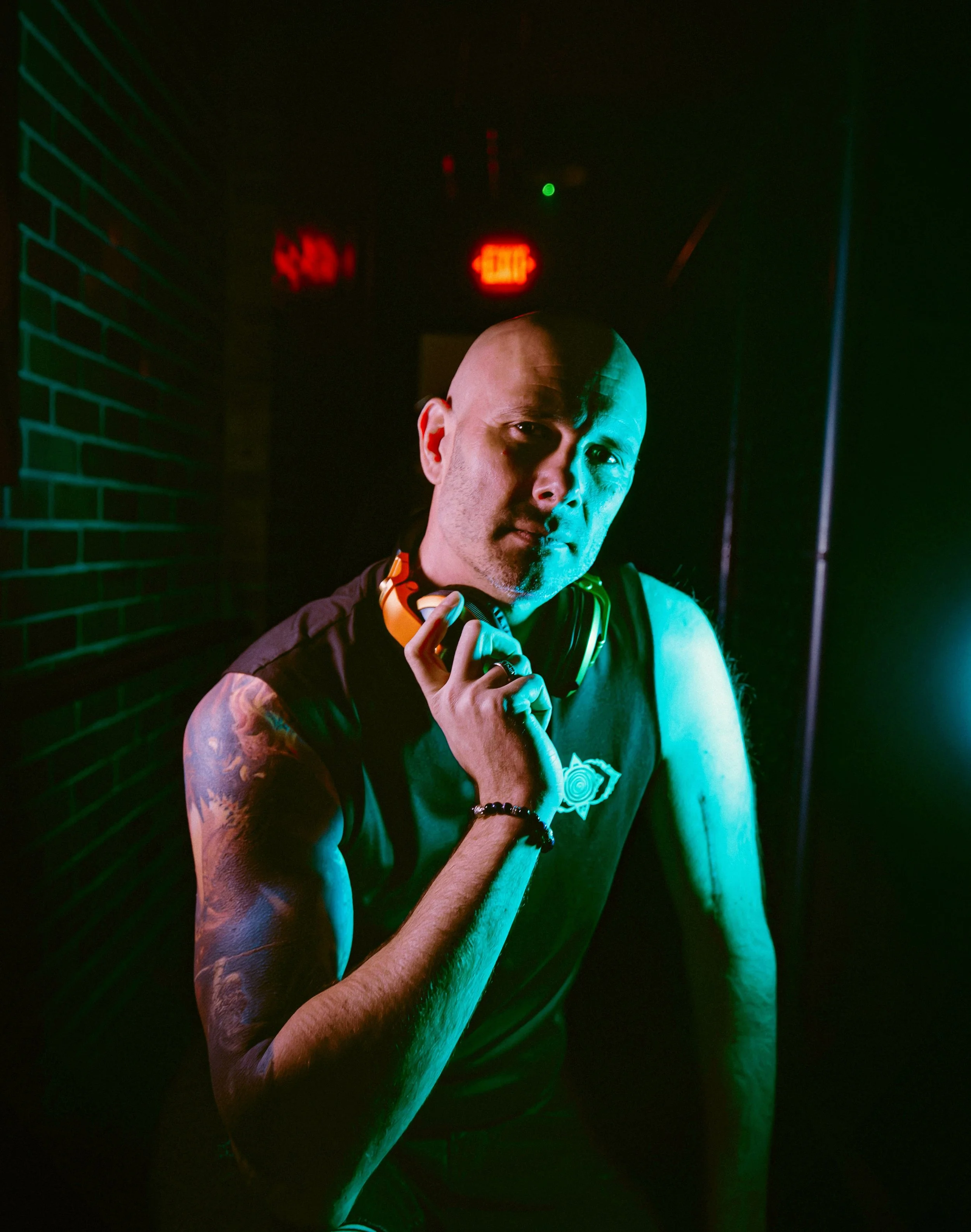 A man with a shaved head and tattoos on his right arm poses in colored lighting in front of a brick wall and exit signs.