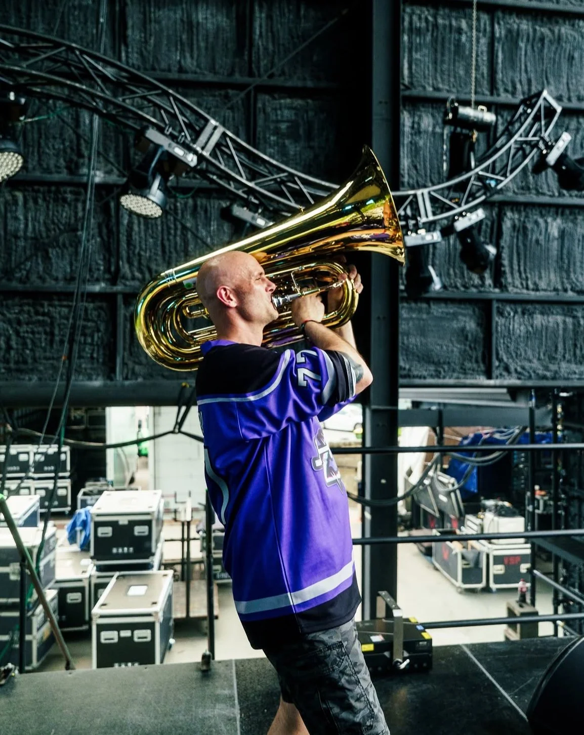 A man wearing a purple sports jersey is playing a large brass tuba on a stage backstage area with equipment and lighting setup.