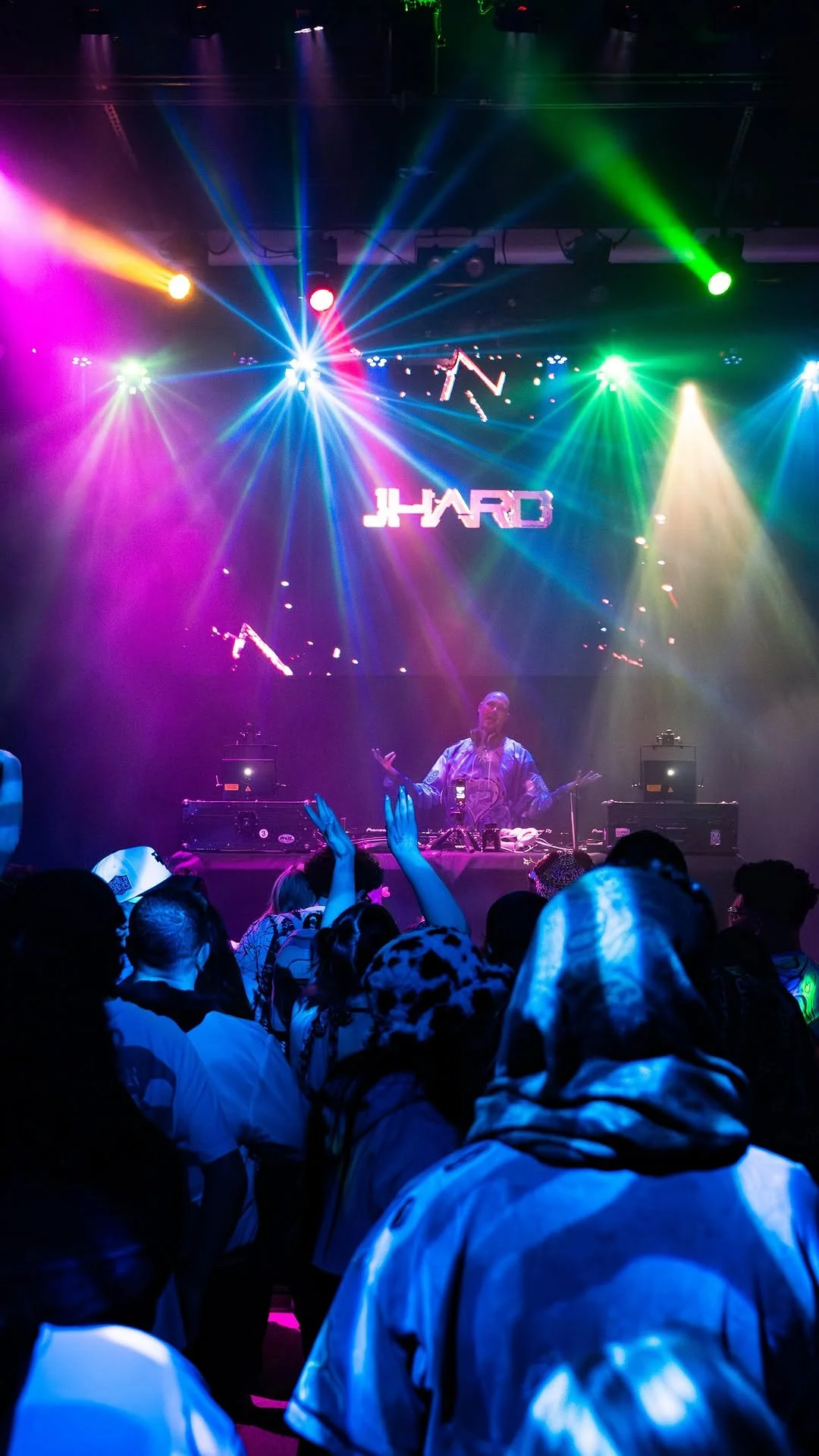 A DJ performs on stage at a concert with colorful rainbow lights shining down. The crowd is dancing and raising their hands.