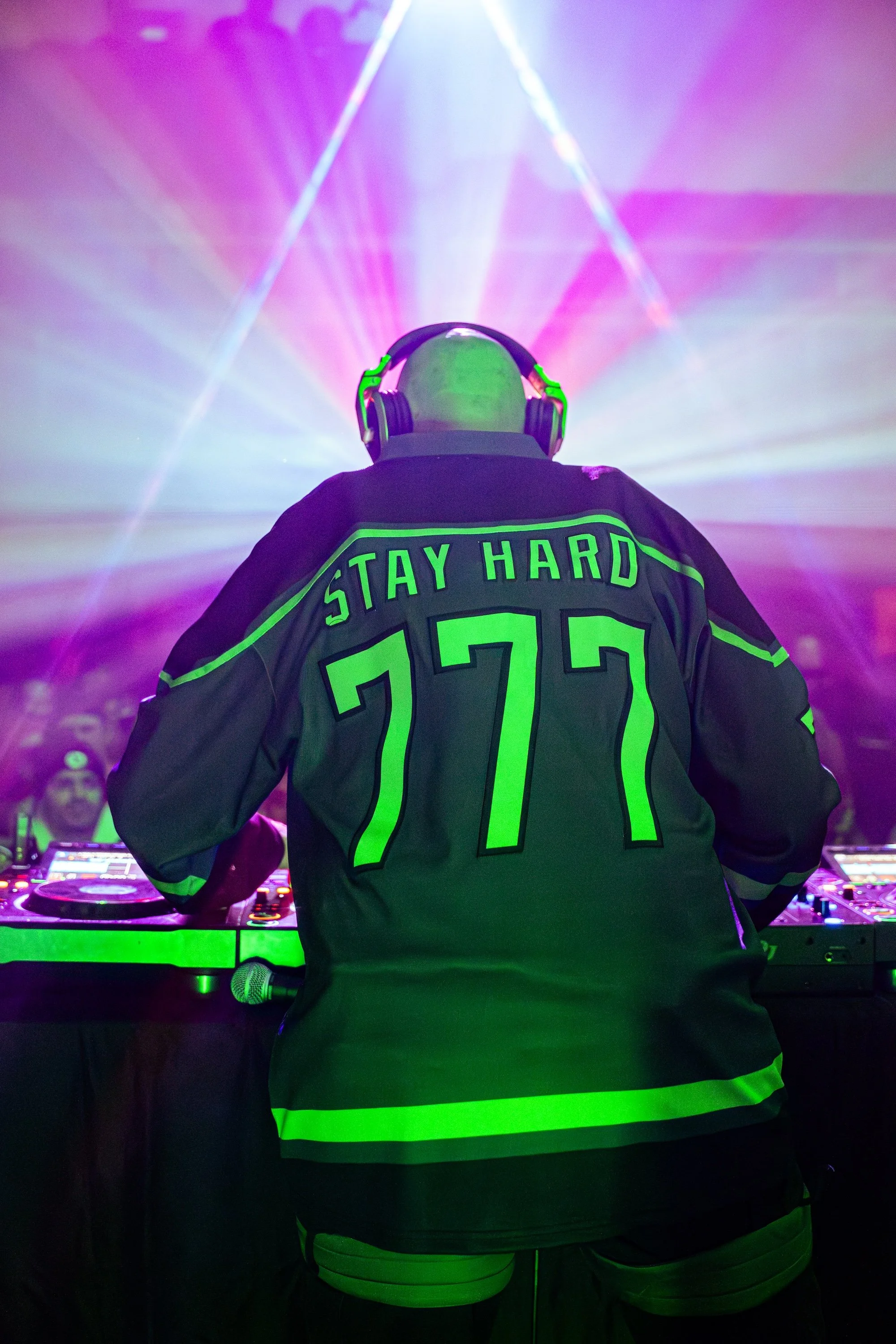 DJ performing at a club with colorful laser lights, wearing a black jacket with bright green accents and the words 'STAY HARD' and the number '725' on the back.