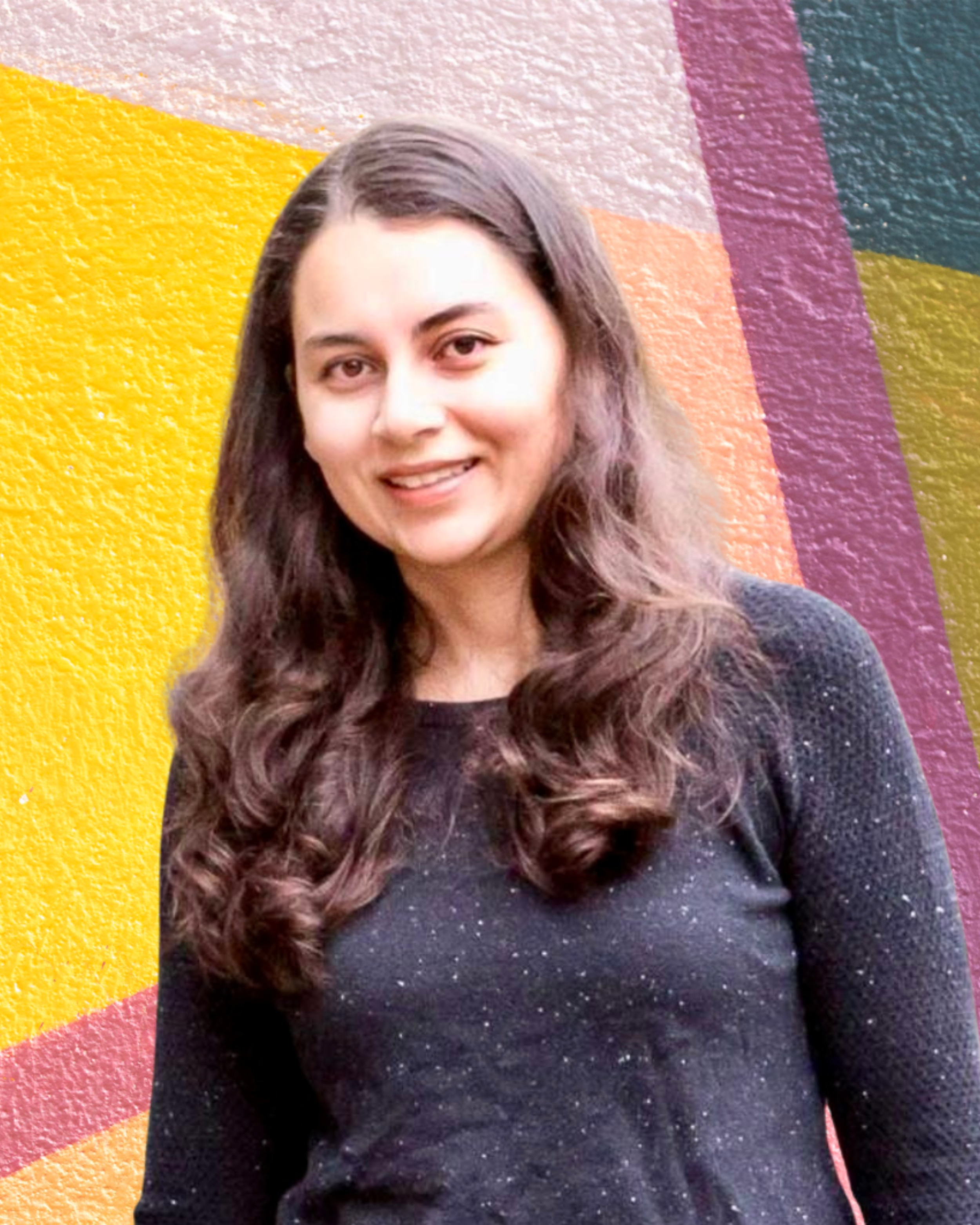 A woman with long wavy hair smiling in front of a colorful abstract painted wall.