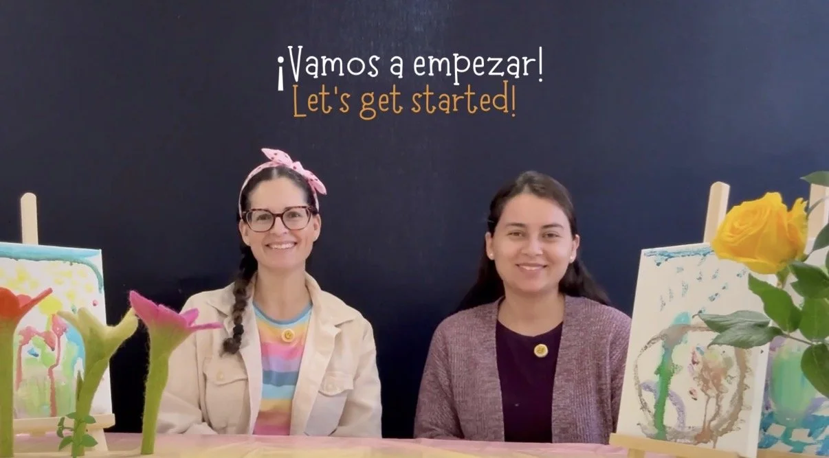 Two women sitting at a table with colorful artwork and flowers, smiling at camera, with a blackboard in the background that says "Let's get started!" in Spanish and English.